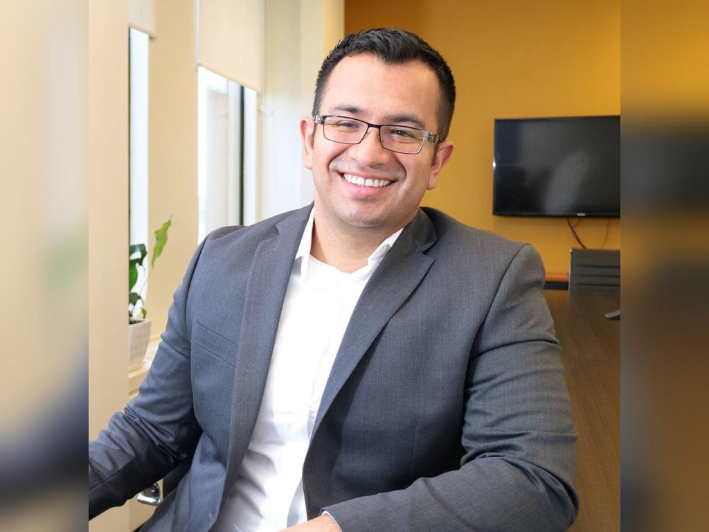 Juan Olarte is a man with dark hair and wire rimmed glasses. He is seated and smiling directly into the camera.