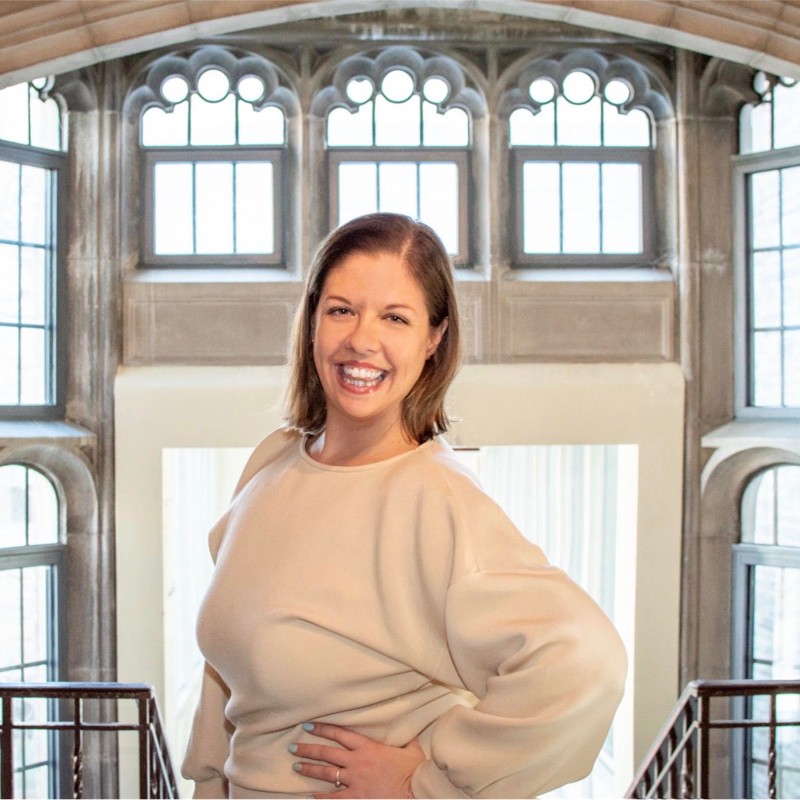 Kate Brown. A woman with shoulder length brown hair, smiling and standing in front of a stained glass window.
