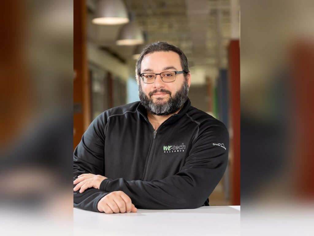 John-Marc is a gentleman with short, dark hair, glasses and a neat beard flecked with white. He's wearing a sweater branded with the WEtech Alliance logo.