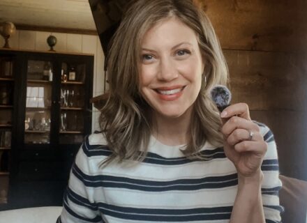 Christine Briggs is a woman with shoulder length blond hair looking into the camera. She's wearing a striped top and holding a mic.